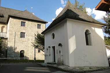 Historical view of a small old chapel with a white facade and a slate roof, in the foreground a street. | © Zell am See-Kaprun Tourismus
