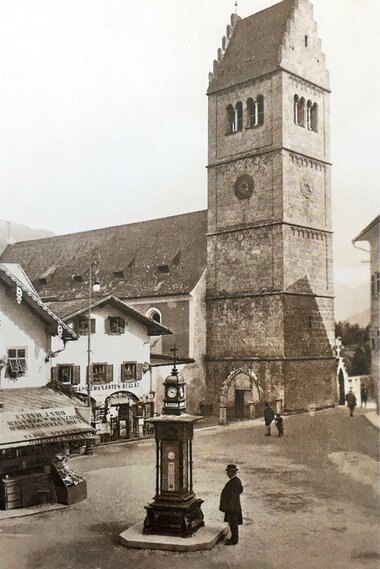 Historical city scene with an old church tower, clock shop, and people in public space.