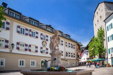 Historical fountain in a city with old buildings, people, and outdoor tables on a sunny day. | © Zell am See-Kaprun Tourismus