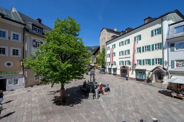 View of a bustling square with a large tree, seating areas, and historic buildings in the background. | © Zell am See-Kaprun Tourismus