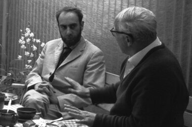 Two people sitting at a table engaged in conversation, with various bowls and small plates on the table.