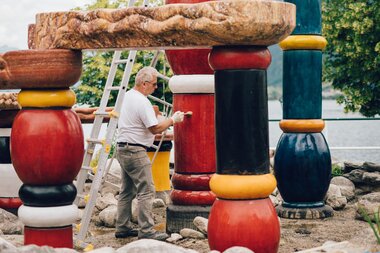 A man works on a large, colorful outdoor art piece made of big, painted wooden pillars. | © Zell am See-Kaprun Tourismus
