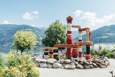 Colorful artistic water fountain with red, black, white, and brown pipes on a paved area, set against a natural backdrop with mountains. | © Zell am See-Kaprun Tourismus