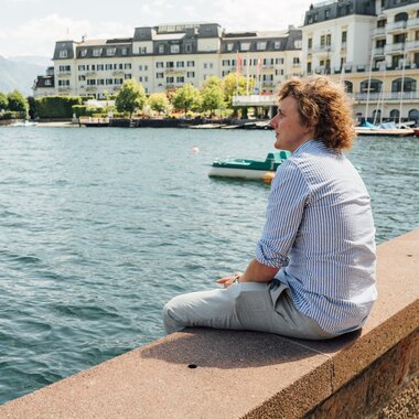 Person sitting relaxed by a lakeside, overlooking the water and cityscape in the background. | © Zell am See-Kaprun Tourismus