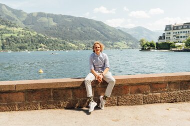 Person sitting on a stone wall by the lake with mountains in the background, sunny day | © Zell am See-Kaprun Tourismus