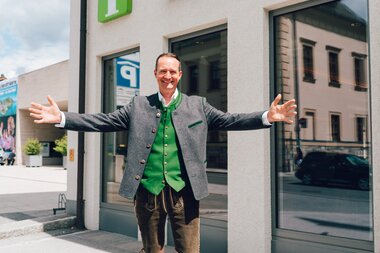 A tourist in traditional Austrian attire stands in front of the Zell am See Kaprun visitor center with open arms. | © Zell am See-Kaprun Tourismus
