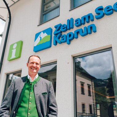 Staff member outside the tourist information center in Zell am See-Kaprun with blue and white signage and a green info symbol. | © Zell am See-Kaprun Tourismus