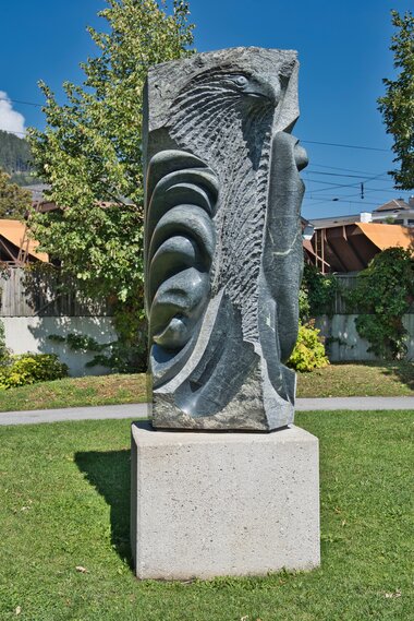 Stone sculpture featuring an eagle's head and abstract shapes on a pedestal in a park.