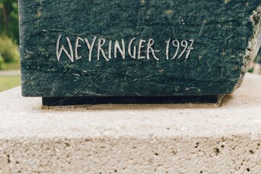 Inscription on a sculpture: 'Weyringer 1997' in white letters on a dark surface.