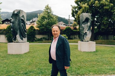 A man standing in a park with sculptures in the background, surrounded by trees and hills.