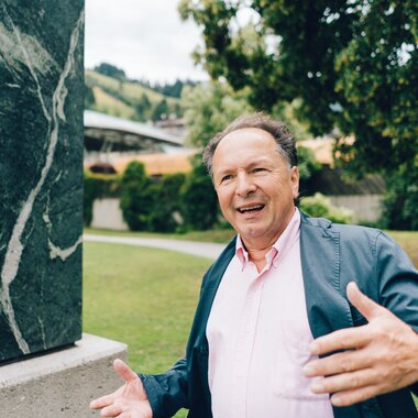 A smiling person speaking next to a green marble sculpture outdoors, surrounded by trees and a well-kept lawn.