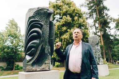 A person stands in front of a modern stone sculpture in a park with trees.