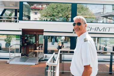 Captain stands on a ship, wearing a white uniform with captain insignia and sunglasses.