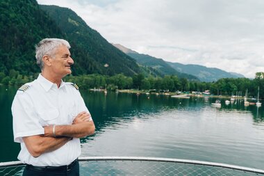 A captain stands on a boat overlooking a calm lake surrounded by mountains and lush greenery.