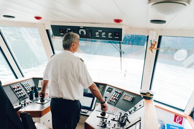 Captain standing in the ship's control room, operating the controls and looking out the large windows at the water.