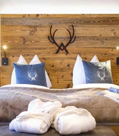Cozy bedroom with wooden panel wall, deer head decor, bed with white and blue pillows, towels, and a magazine on the bed's edge. | © Nikolaus Faistauer Photography
