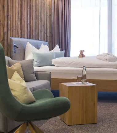 Cozy hotel room with a bed, an armchair, a sofa, and a small wooden table, in front of a window with curtains. | © Nikolaus Faistauer Photography