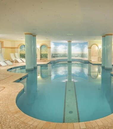Indoor view of a wellness or thermal bath area with a large, oval pool and lounge chairs in the background. | © Nikolaus Faistauer Photography