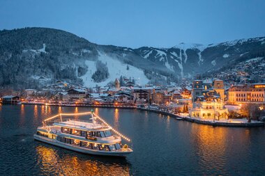 Night view of a lit-up boat on a river in a snowy mountain town. | © Nikolaus Faistauer Photography