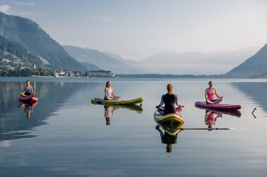 Four people practicing yoga on paddleboards on a calm lake in Zell am See with mountains in the background. | © Zell am See-Kaprun Tourismus