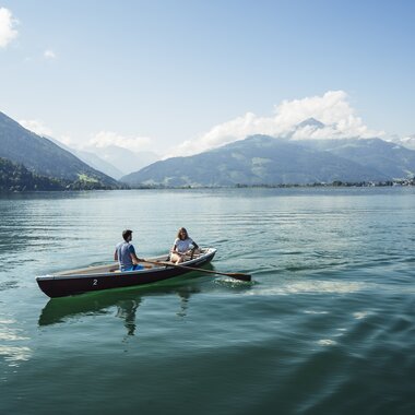 Two people paddling a canoe on a calm lake, surrounded by mountains under a blue sky. | © Zell am See-Kaprun Tourismus