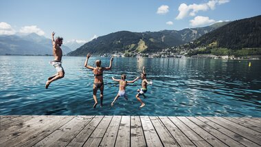 Children jump and splash in Lake Zeller on a sunny day, surrounded by mountains and a small town in the background. | © Korbinian Seifert