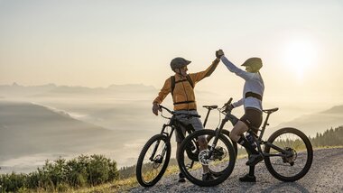 Two people mountain biking on a hill at sunrise, exchanging a high five or celebration gesture. | © Johannes Radlwimmer