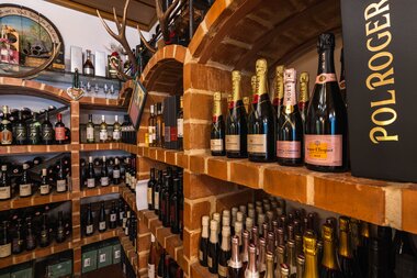 Shelf with bottles of champagne, wine, and spirits in a gourmet shop, brick shelves, cozy atmosphere. | © Johannes Radlwimmer