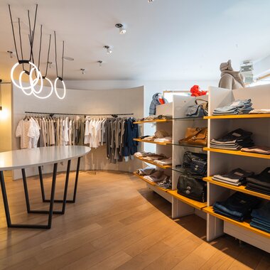Interior of a clothing store with racks of clothes, shelves with folded apparel, and modern lighting. | © Johannes Radlwimmer