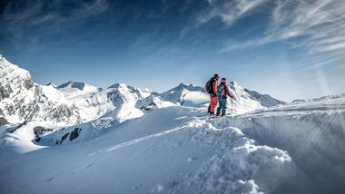 Two skiers on a snowy mountain ridge with peaks in the background and a clear sky. | © Kitzsteinhorn