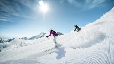 Two skiers on a groomed slope at Kitzsteinhorn under sunshine and a clear blue sky. | © Kitzsteinhorn