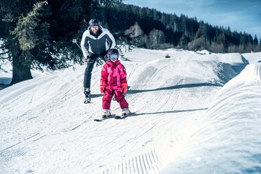 An adult and a child skiing on a snowy slope, surrounded by snow and trees, on a sunny day. | © Kitzsteinhorn