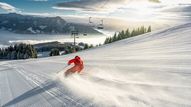 Skier skier descending a snowy slope at sunrise with a mountain landscape, ski lift, and pine trees in the background. | © Zell am See-Kaprun Tourismus