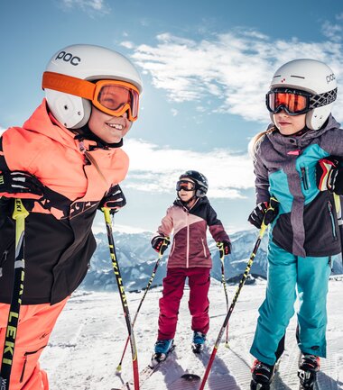 Children skiing on the Schmittenhöhe, smiling and dressed in winter sports gear, in the snow with mountains in the background. | © Zell am See-Kaprun Tourismus