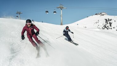 Two people skiing down a snowy slope under a clear blue sky, with ski lifts in the background. | © Zell am See-Kaprun Tourismus