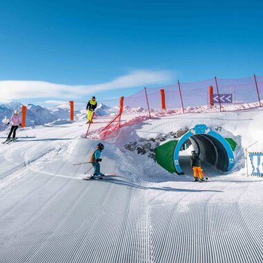Children and teenagers skiing on a snow-covered slope with a tunnel attraction and mountain scenery. | © Schmittenhöhe