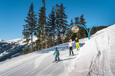 Skiers and children on the Schmittenhöhe in the ski area, with mountains and pine trees in the background, on a clear winter day. | © Schmittenhöhe