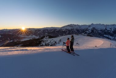 Skier on a snowy mountain during sunset with a mountain range in the background. | © Schmittenhöhe