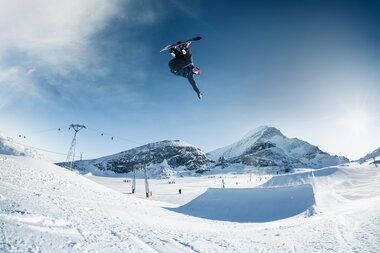 Snowboarder performing a jump in a snowpark with snow-covered mountains in the background. | © Markus Rohrbacher