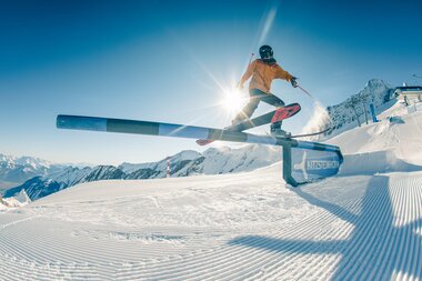 Skier skier jumps over a snow platform in a snow park with mountains in the background under sunshine. | © Markus Rohrbach