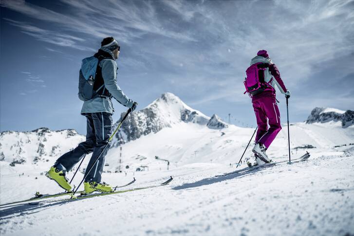 Two skiers on a snowy slope in front of a glacier with mountains in the background. | © Kitzsteinhorn