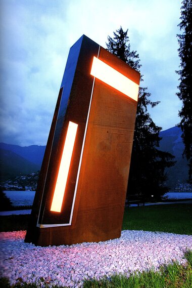 Modern light sculpture with geometric shapes, illuminated at dusk, in a park with trees and mountains in the background