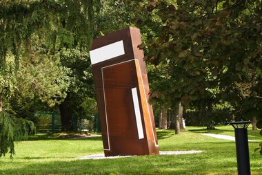 Large modern sculpture made of metal and wood in a park with trees and grass.