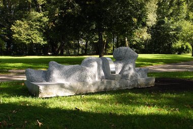 Impressive sculpture made of light-colored stone in a park, surrounded by trees and sunlit grass.