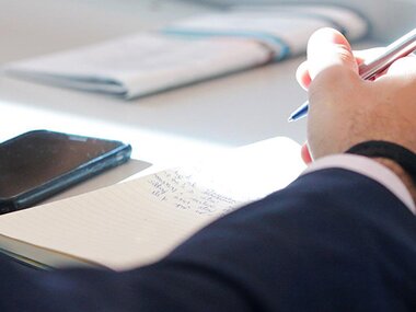 Close-up of a person writing at a desk with a notebook, smartphone, and documents in the background.