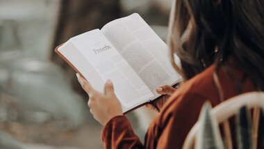 Person reading an open book outdoors, focusing on hands and the book.