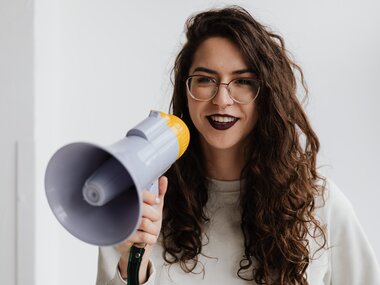 A person is holding a megaphone, wearing glasses, with dark, curly hair.