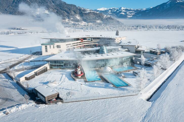 Aerial view of the wintery Tauerna Spa thermal baths with water slides and steam clouds, surrounded by snow and mountains. | © TAUERN SPA Zell am See-Kaprun