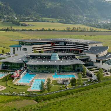 Modern spa resort with outdoor pools surrounded by green fields and mountains in the background. AI-generated text. | © TAUERN SPA Zell am See-Kaprun