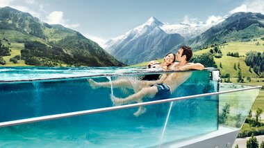 Two people relax in an infinity pool with mountain views in a spa setting. | © TAUERN SPA Zell am See-Kaprun